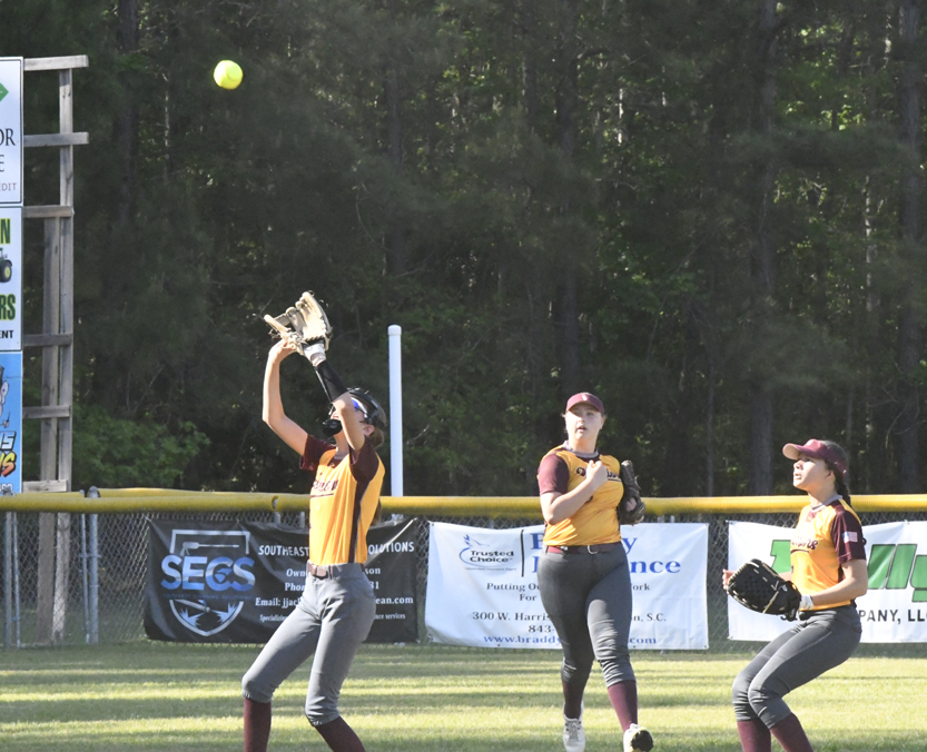 Three softball players in yellow uniforms prepare to catch a ball in the outfield, with trees and advertising banners in the background.