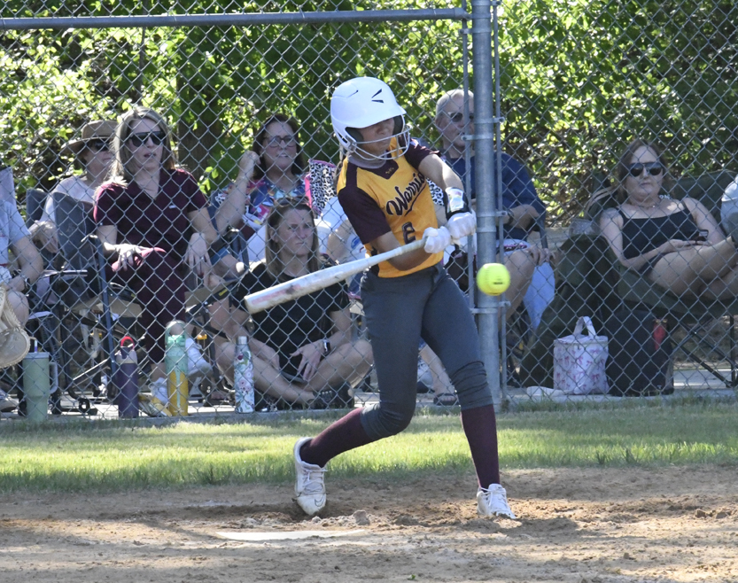 A softball player in a white helmet swings at a pitched ball, while spectators watch from behind a chain-link fence.