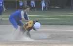 A softball player in yellow slides into base while a player in blue attempts to tag them, dust rising from the ground.