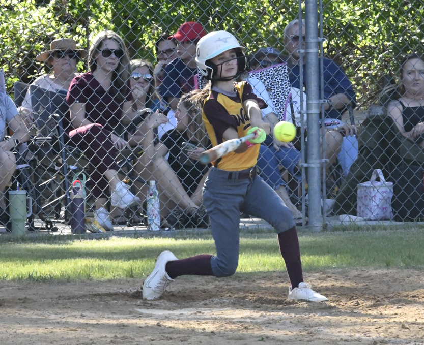A softball player in a helmet swings at a pitch, while spectators watch from behind a chain-link fence on a sunny day.