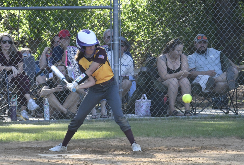 A softball player in a yellow and black uniform swings at a pitched ball, with spectators watching behind a chain-link fence.