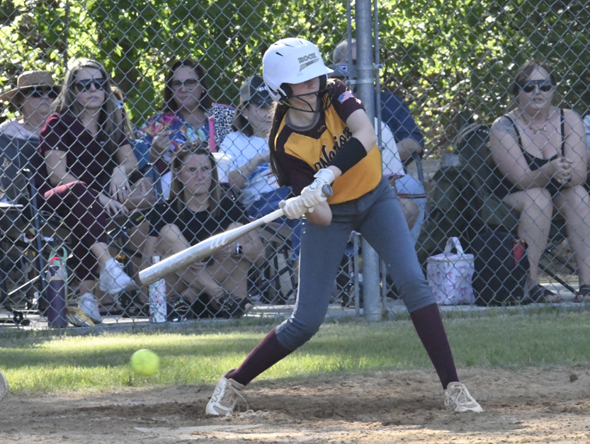 A softball player in a yellow and maroon uniform swings at a pitch during a game, with spectators watching from behind a chain-link fence.