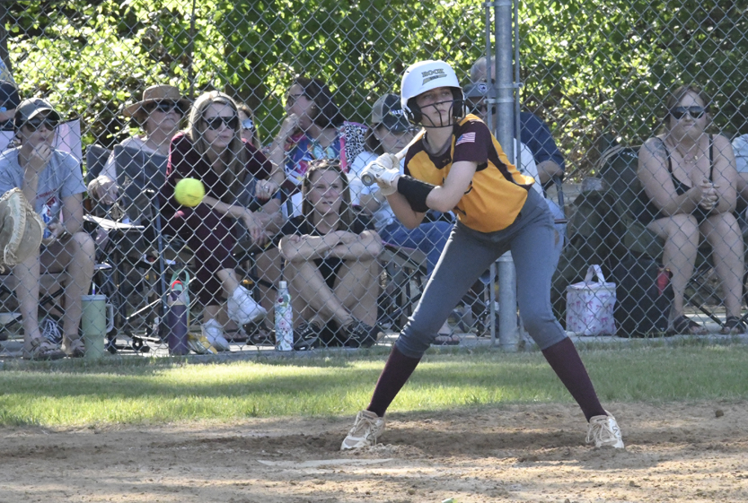 A softball player in a yellow and maroon uniform prepares to bat as the ball approaches, with spectators watching behind a chain-link fence.