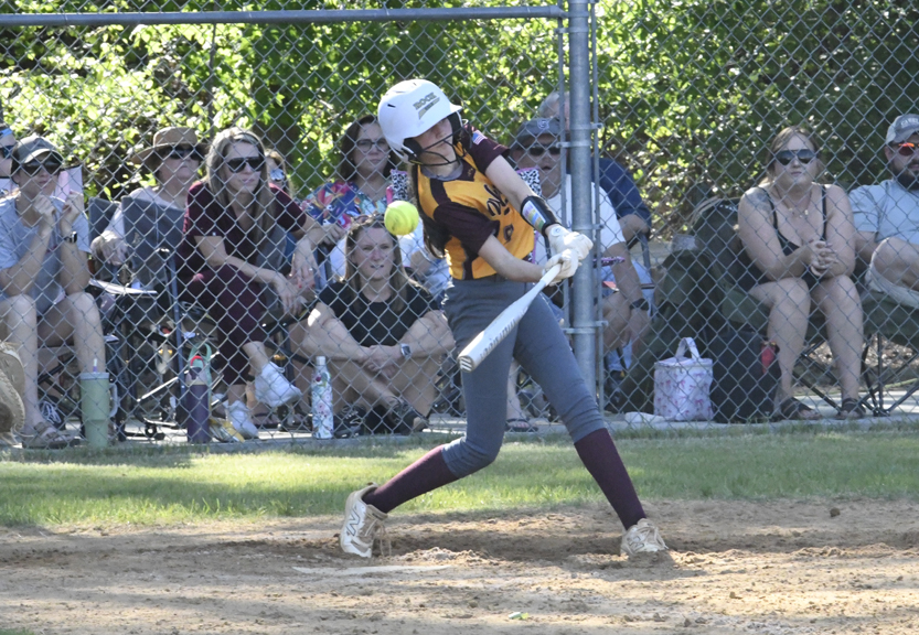 A softball player in uniform swings a bat at a pitch, with spectators watching from behind a chain-link fence on a sunny day.