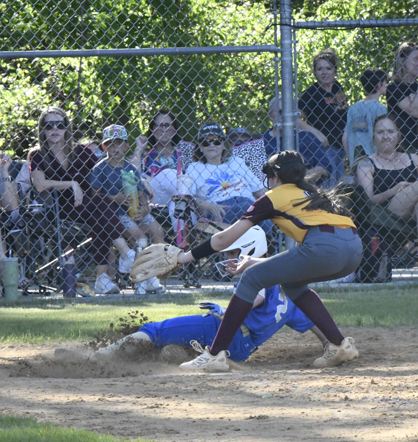 A softball player in blue slides into home plate as a player in yellow attempts to tag, with spectators watching from behind a fence.