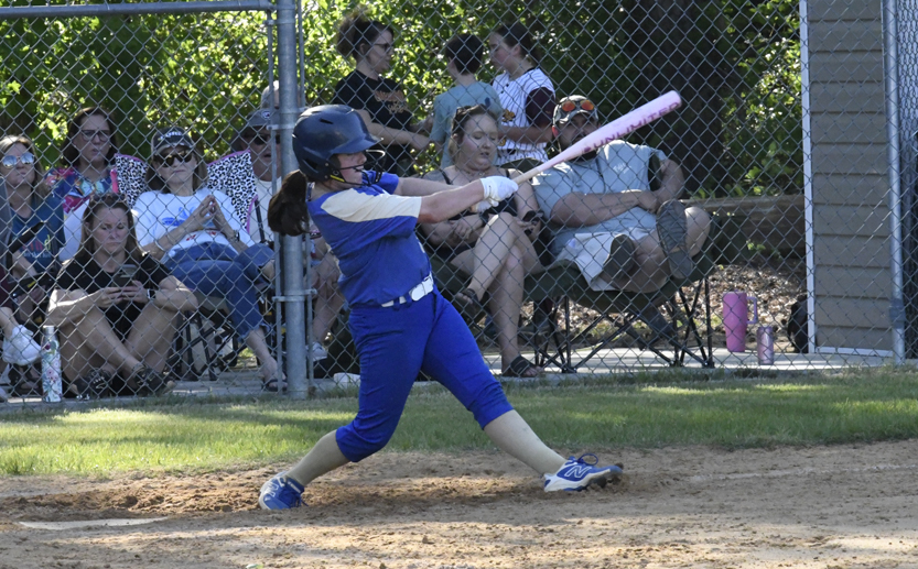 A softball player in blue uniform swings a bat during a game, with spectators seated behind a chain-link fence in the background.