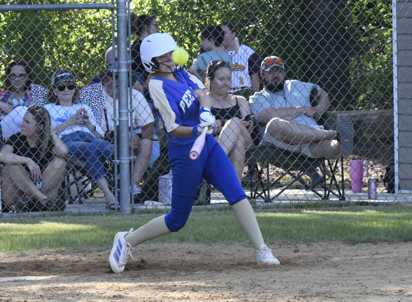A softball player in blue uniform gets hit in the face by a ball while batting, spectators watch from behind a fence.