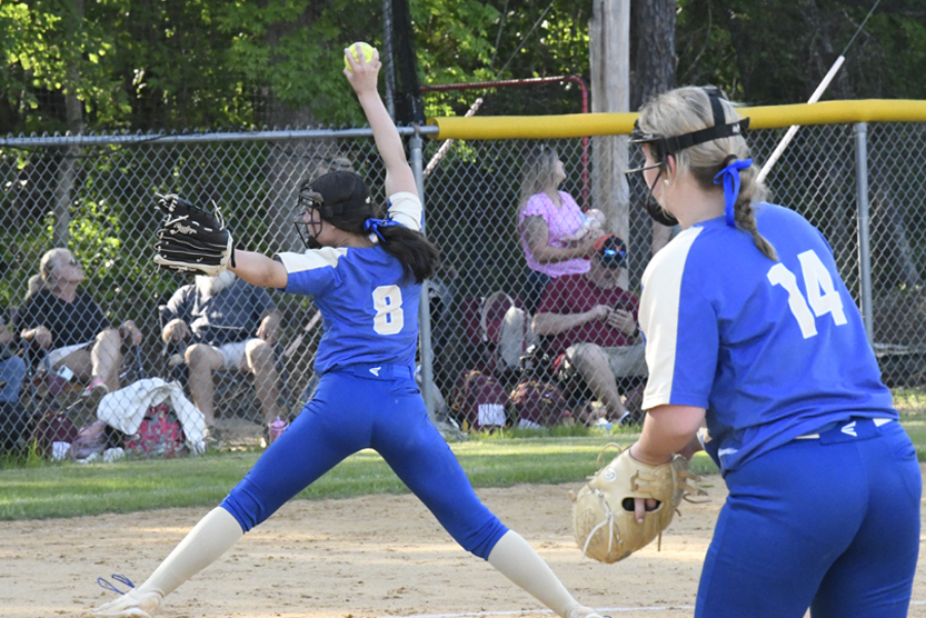 A softball pitcher in blue uniform winds up to throw, while a teammate stands ready. Spectators watch from behind a fence in the background.