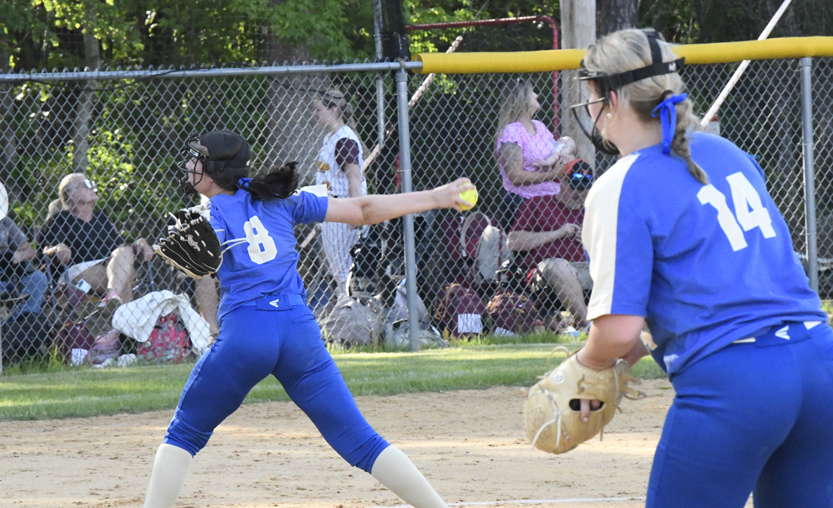 A softball pitcher in blue winds up to throw the ball, while another player prepares to catch. Spectators watch from behind a chain-link fence.