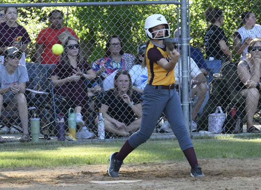 A softball player in a yellow and brown uniform swings at a pitch, with spectators watching behind a chain-link fence.