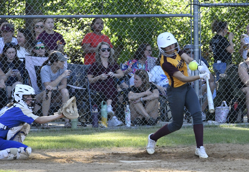 A softball player wearing a helmet swings at a pitch during a game, while spectators watch from behind a chain-link fence in the background.