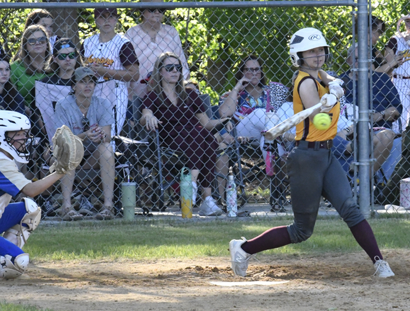 A softball player in a yellow jersey swings a bat, making contact with the ball, while spectators watch from behind a chain-link fence.
