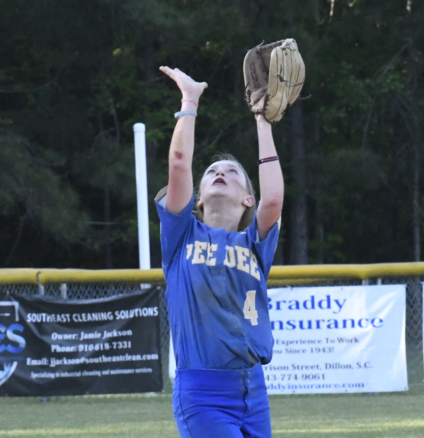 A softball player in a blue uniform is reaching up with a glove to catch a ball during a game on an outdoor field.