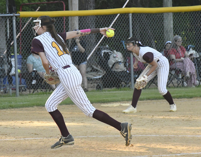 A softball pitcher throws the ball during a game, with a teammate ready in the infield and spectators watching from behind the fence.