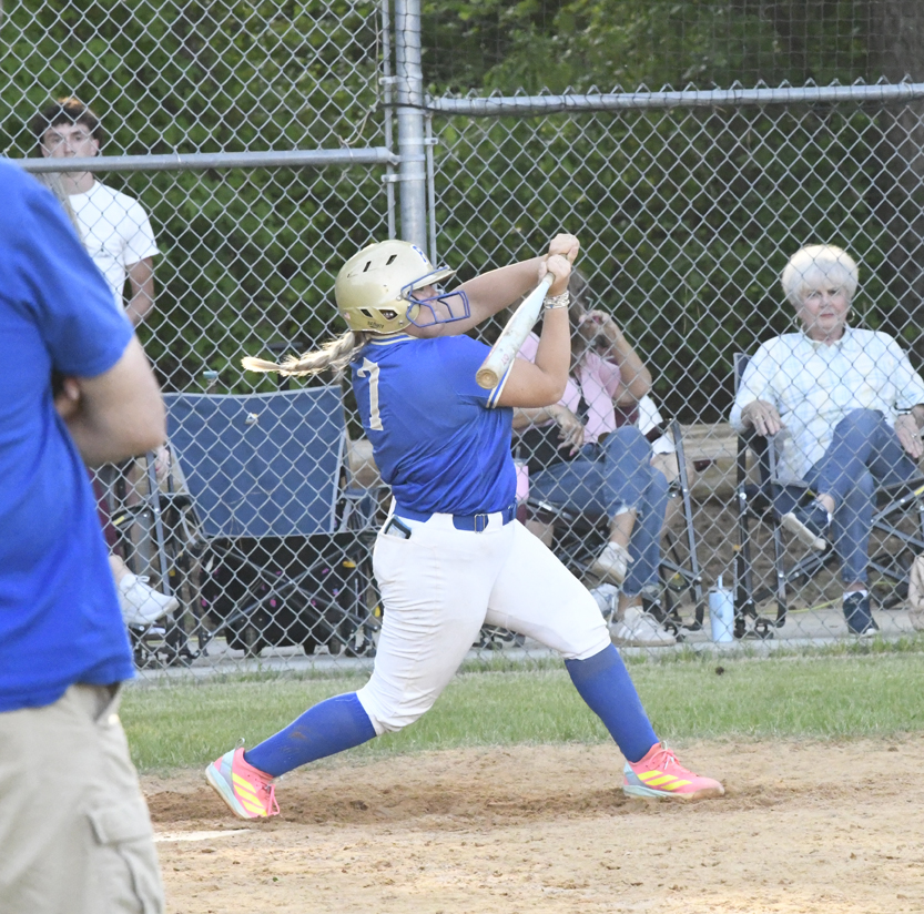 A softball player in blue swings a bat during a game, with spectators seated behind a chain-link fence in the background.