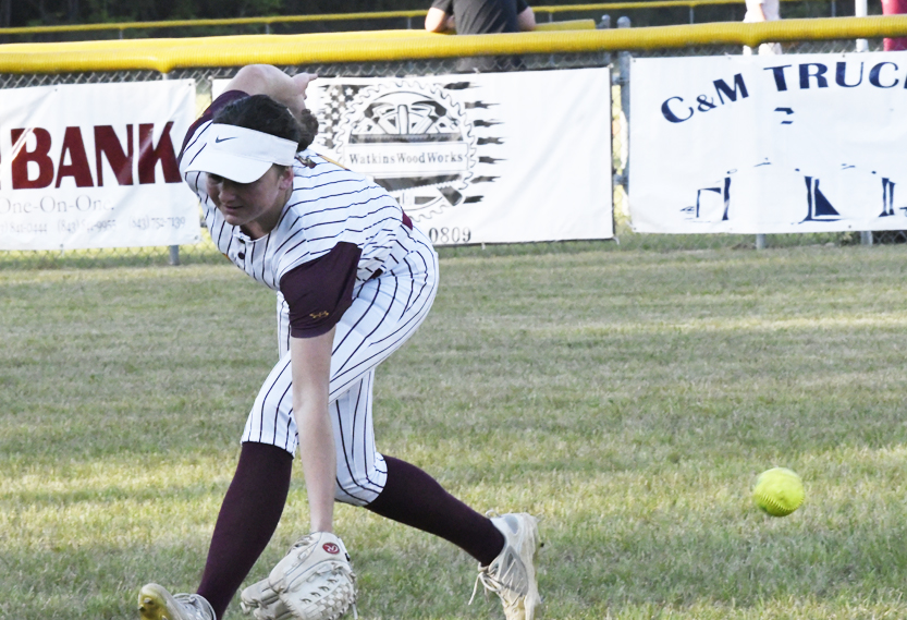 A softball player in a striped uniform bends down to field a ground ball on a grassy field during a game.