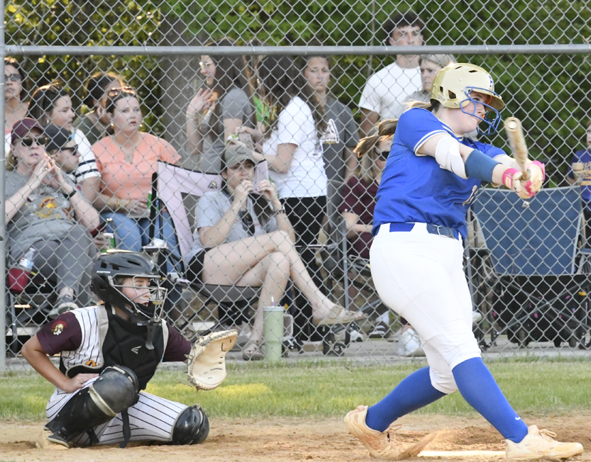A softball player in blue uniform swings at a pitch as the catcher and spectators watch from behind a chain-link fence.