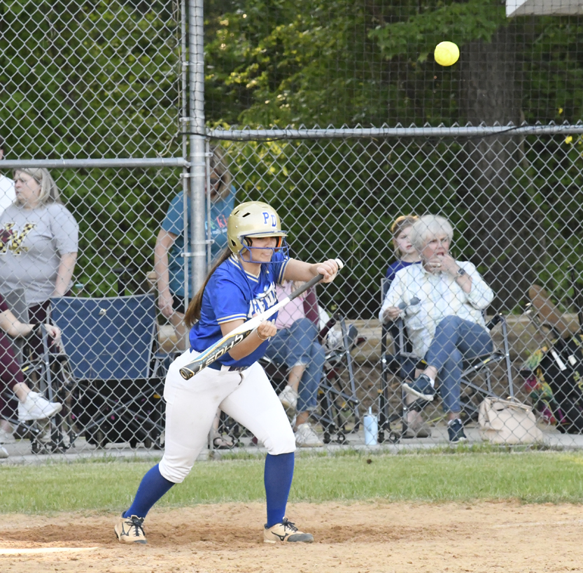 Softball player in blue uniform prepares to bunt as the ball approaches, with spectators seated behind a chain-link fence in the background.