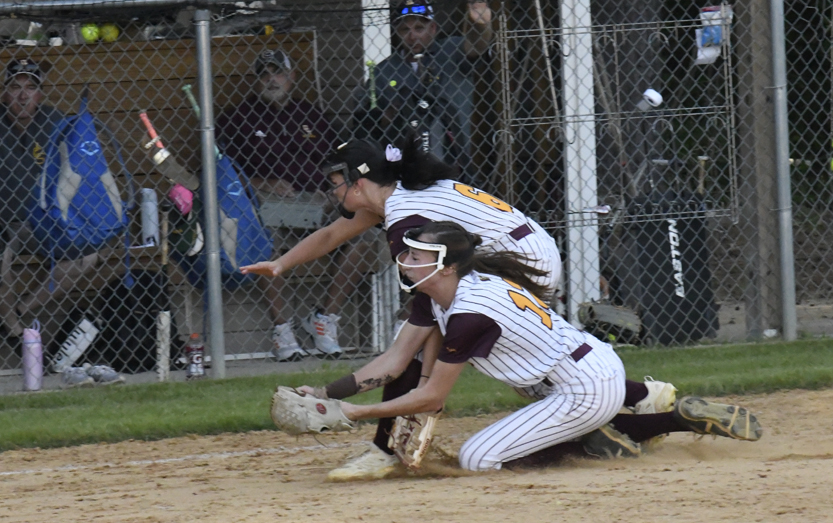 Two softball players in striped uniforms attempt to tag a runner sliding into base during a game, with a chain-link fence and spectators in the background.