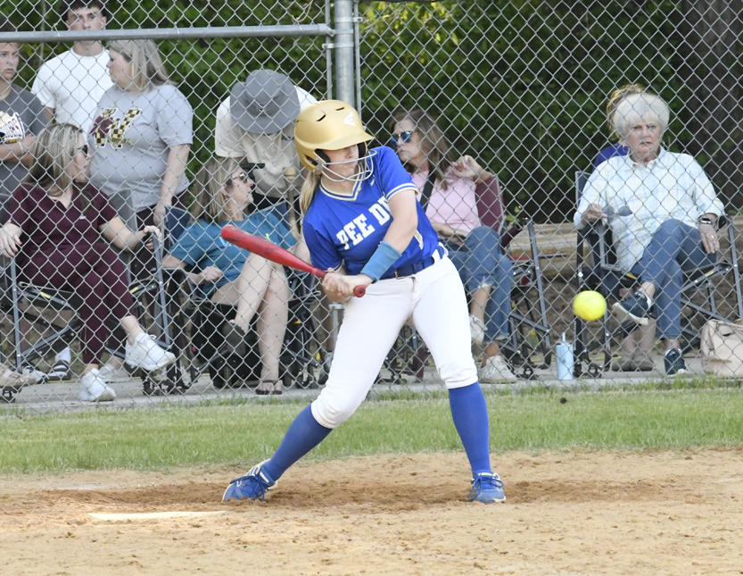 A softball player in blue and white uniform prepares to hit a pitch during a game, with spectators seated behind a chain-link fence in the background.