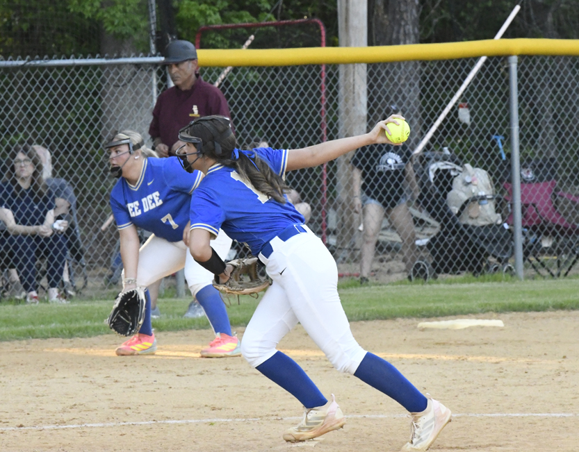 A softball pitcher in blue and white uniform throws a pitch, while another player and an umpire watch on a dusty field.