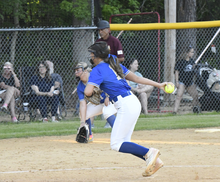 Softball pitcher in blue uniform throws a pitch during a game, with a catcher, umpire, and spectators visible in the background.