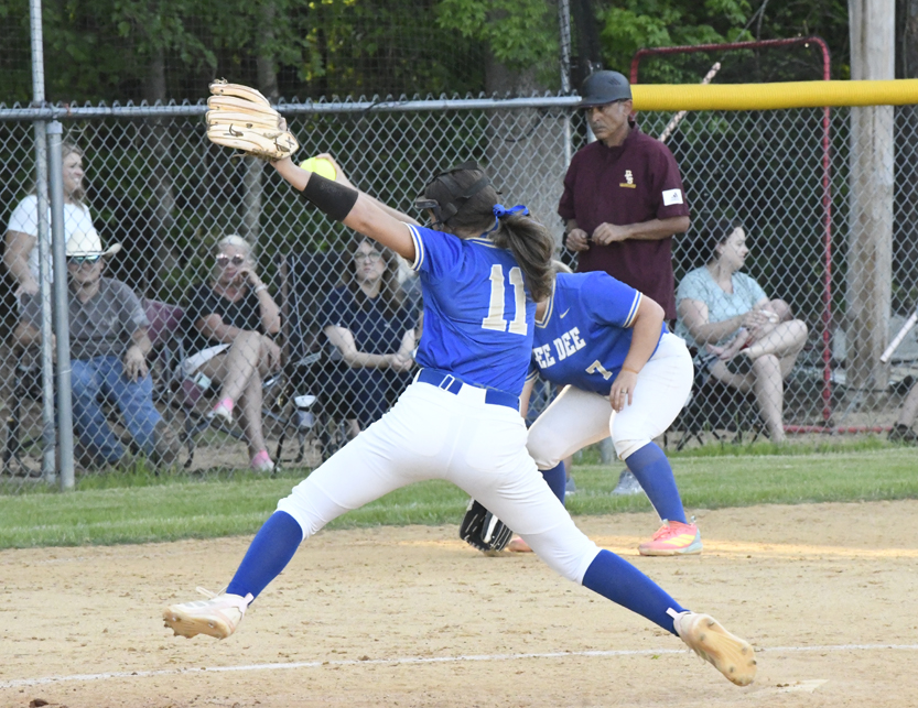 Softball pitcher in blue uniform throws a pitch during a game, with an umpire and spectators visible in the background behind a chain-link fence.