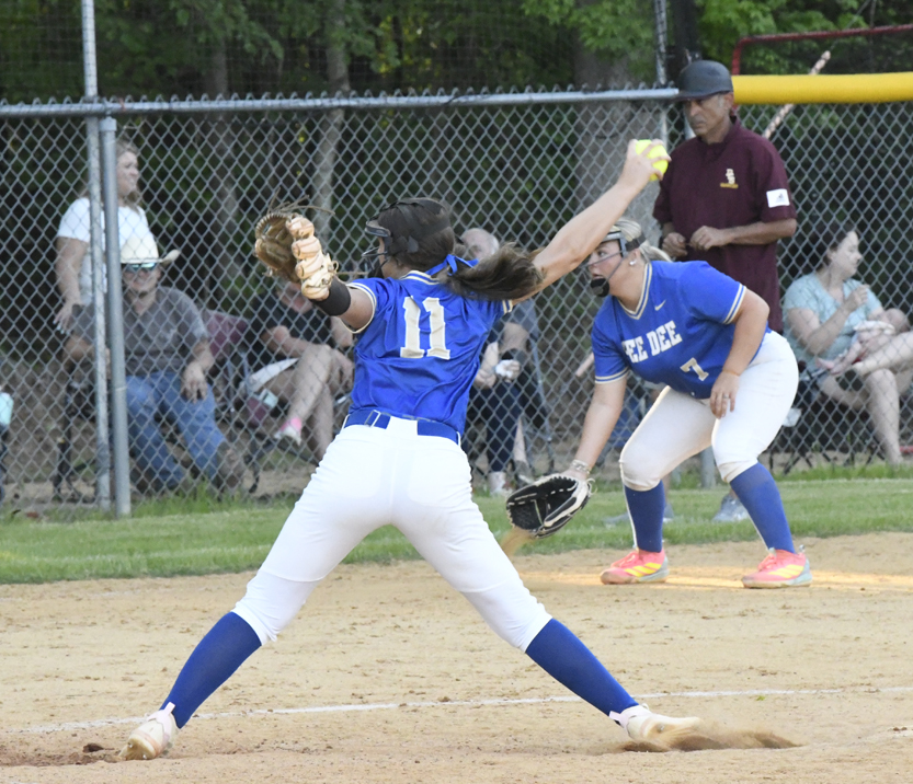 Softball pitcher in blue uniform mid-pitch on dirt field, with teammate ready at base and umpire observing, spectators behind chain-link fence.