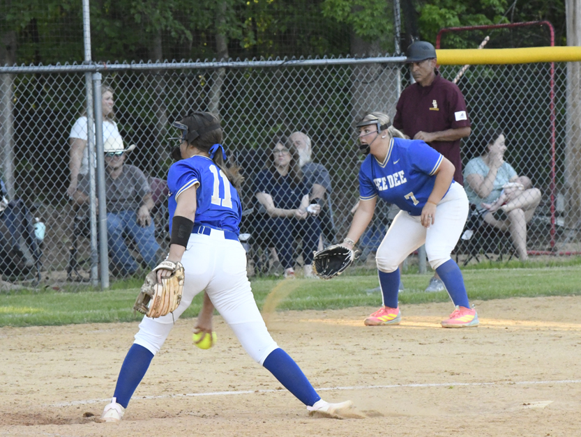 Two female softball players in blue uniforms play on a dirt field, while an umpire and spectators watch from behind a chain-link fence.