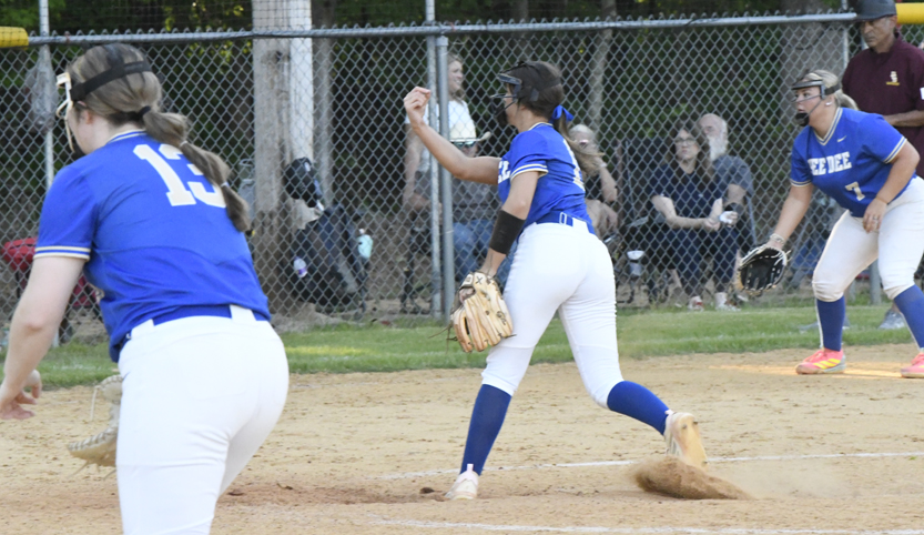Two softball players in blue and white uniforms playing on a dirt field, one preparing to throw the ball. Spectators sit behind a chain-link fence.