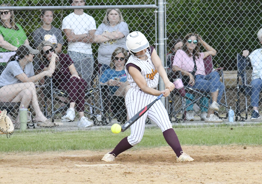 Softball player in striped uniform swinging bat, making contact with ball, with spectators watching behind a fence.