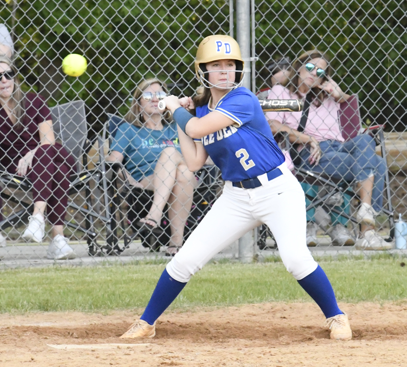 A softball player in blue and white uniform prepares to bat as the ball approaches, with spectators watching behind a chain-link fence.