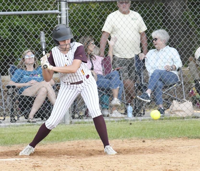 A softball player in uniform prepares to bat, with a crowd of spectators seated behind a chain-link fence in the background.