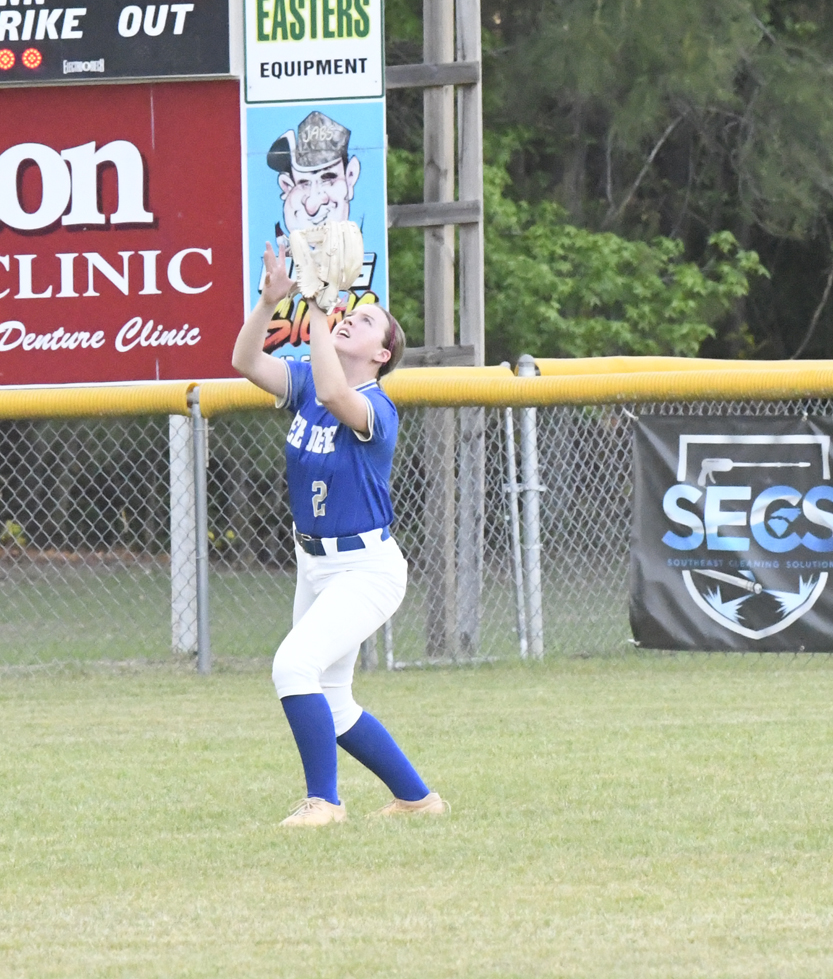 Softball player in blue and white uniform catches a ball in the outfield near a fence and advertising banners.