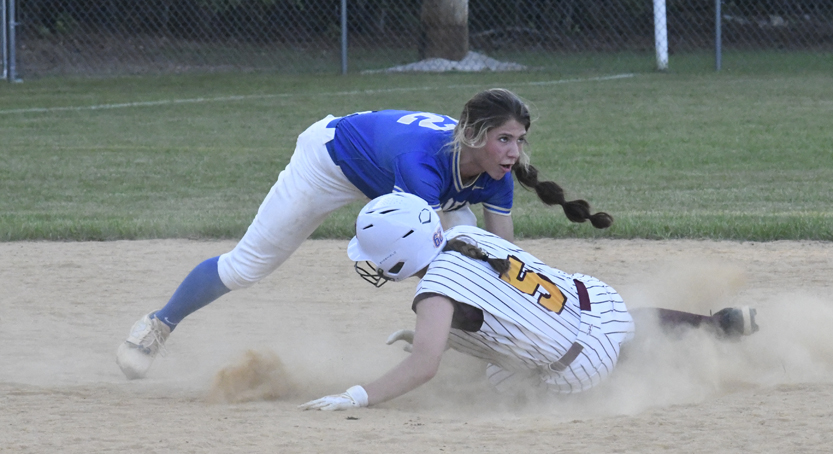 Two softball players in action; one in blue attempts to tag out a sliding player in white uniform and helmet on a dusty infield.