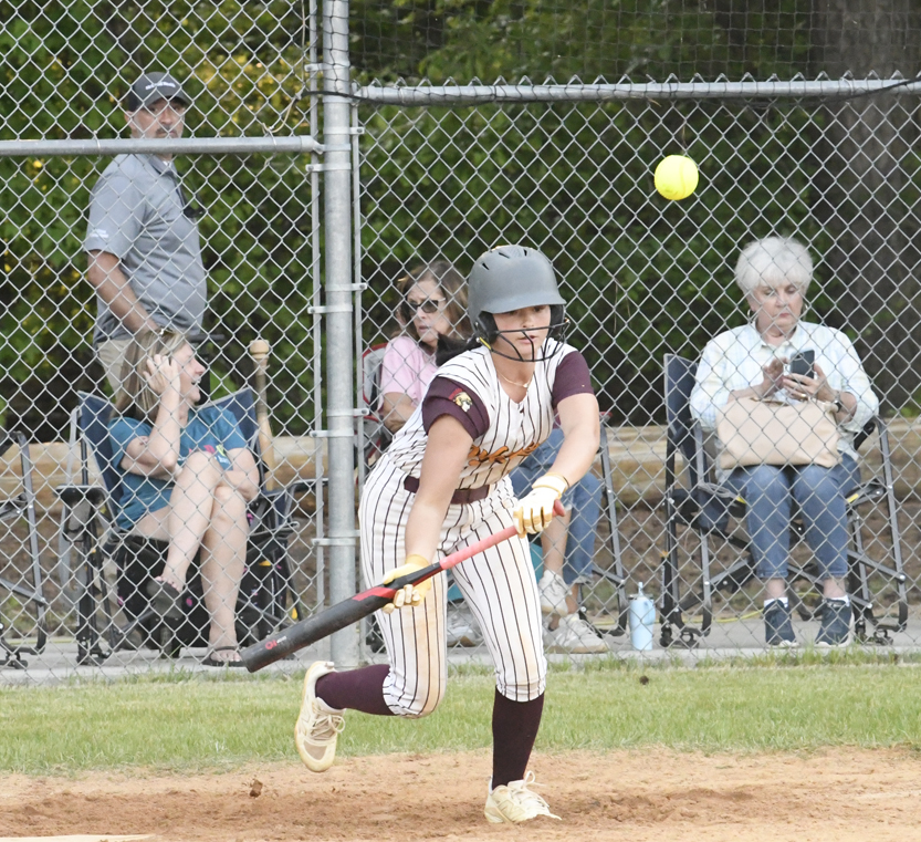 A softball player swings at a pitch on a field, with spectators watching from behind a chain-link fence in the background.
