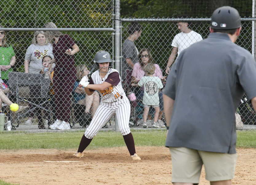 A softball player prepares to bat while wearing a helmet and uniform, with a coach nearby and spectators watching behind a chain-link fence.