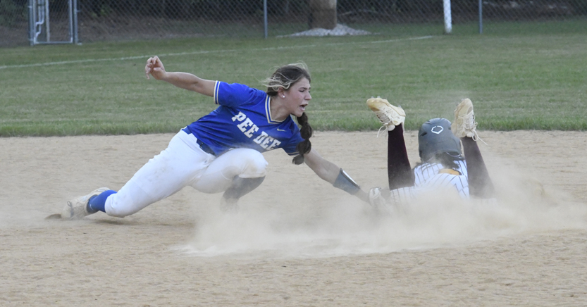 Softball player in blue jersey attempts to tag out a sliding runner at a dusty base during a game on a dirt field.