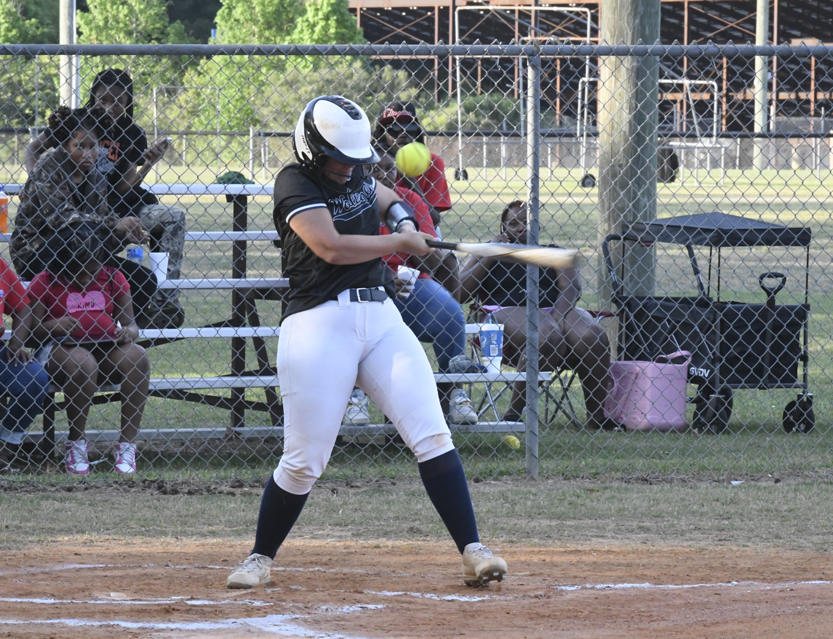 A softball player in a helmet and uniform hits a pitch, with spectators watching from bleachers behind a chain-link fence.