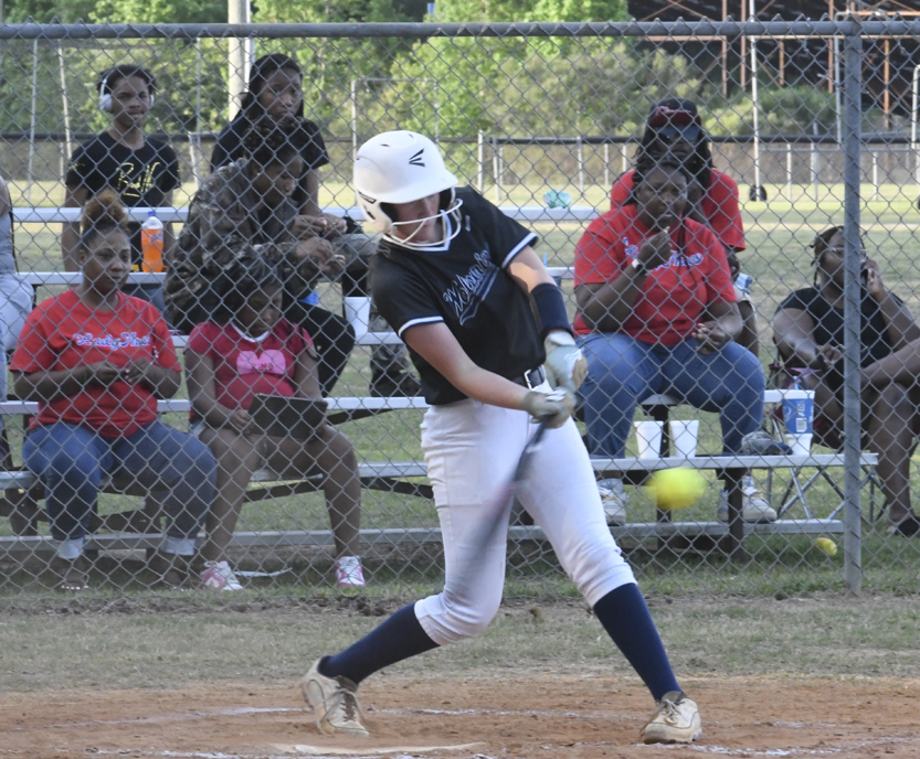 Softball player in uniform hits a yellow softball while spectators watch from metal bleachers behind a chain-link fence at an outdoor field.