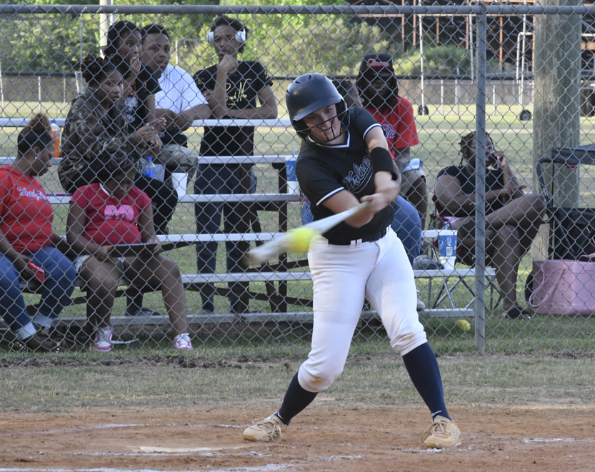 A softball player swings at a pitch during a game, with spectators sitting in bleachers behind a chain-link fence.