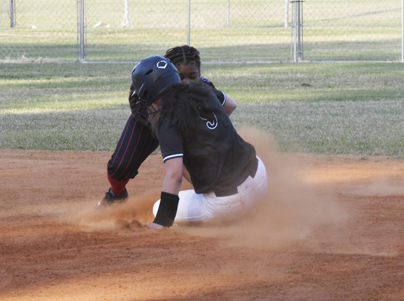 A softball player slides into base as another player attempts to tag them, creating a cloud of dust on the field.