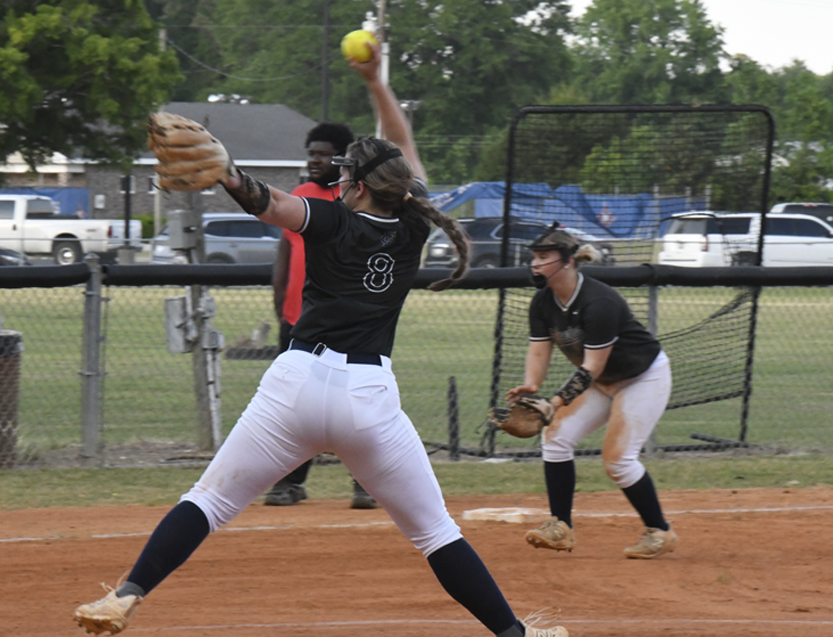 Softball pitcher in mid-throw during a game, with a teammate and an umpire in the background on an outdoor field.