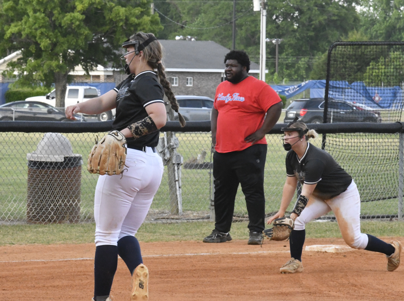 Two softball players in black uniforms are on a field, one pitching and one ready to catch, with a coach observing in the background.