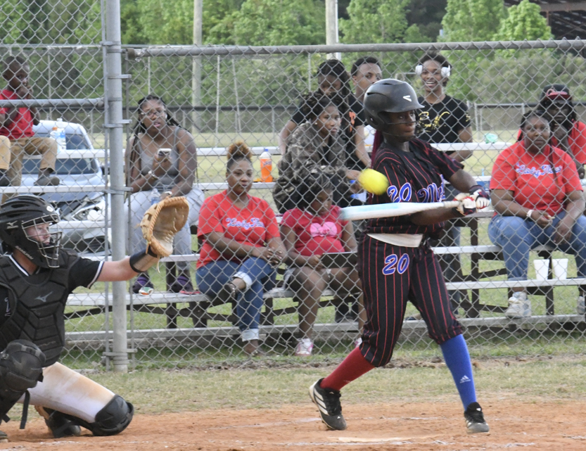 A youth baseball player swings at a pitch while a catcher and spectators watch through a chain-link fence during a game.
