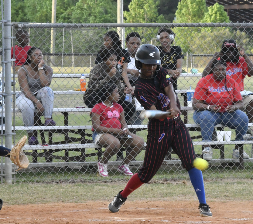 A softball player in a black and red uniform hits a yellow ball as spectators watch from the bleachers behind a chain-link fence.