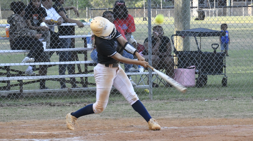 A softball player in a helmet swings a bat at a pitch on a dirt field, with spectators watching behind a chain-link fence.