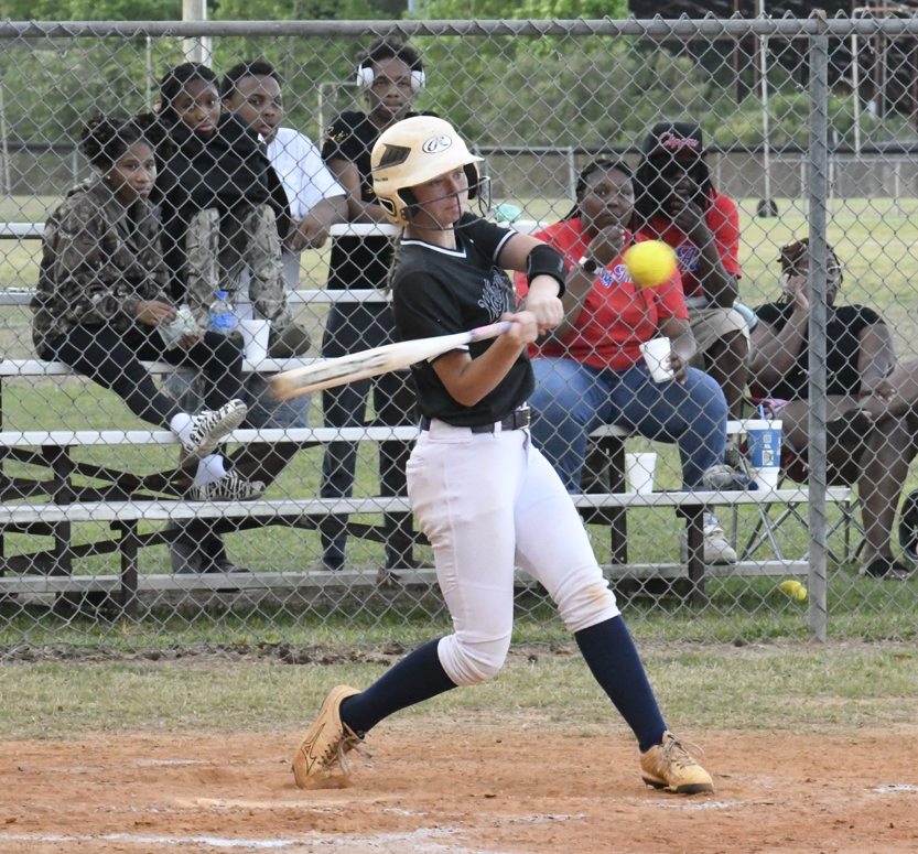 A softball player wearing a helmet swings at a pitch during a game, with spectators watching from bleachers behind a chain-link fence.