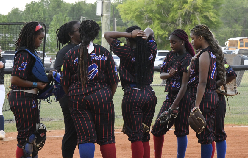 A softball team in black and red uniforms huddles together on the field during a game, listening to their coach.