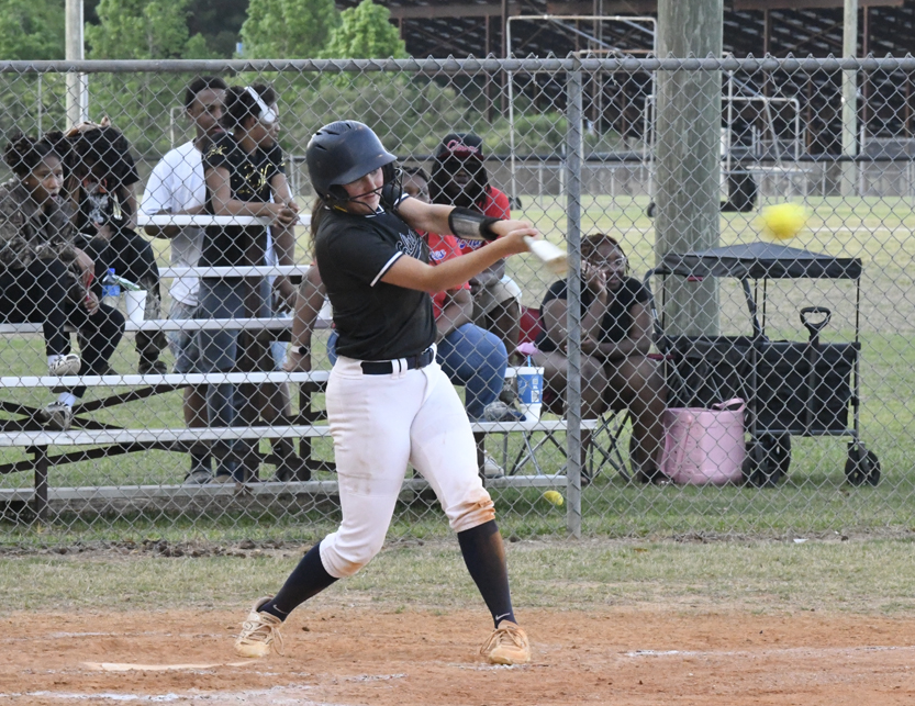 A softball player in a black jersey and white pants hits a yellow ball, with spectators watching from bleachers behind a chain-link fence.